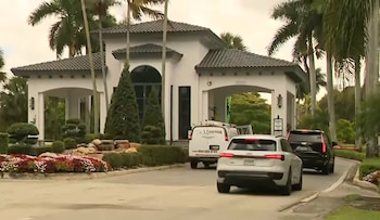 Entrada a una comunidad residencial de lujo con portón blanco, palmeras y jardines floridos. Una van, un SUV blanco y un SUV negro transitan la vía