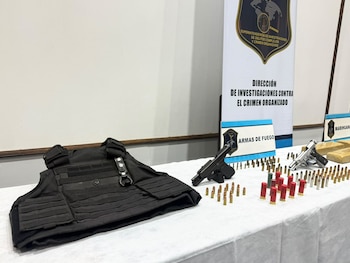 Un chaleco antibalas, dos pistolas, proyectiles de diverso calibre y paquetes rectangulares sobre una mesa blanca frente a paneles policiales