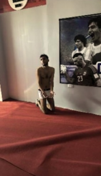Un hombre en ropa interior blanca está arrodillado con las manos juntas sobre una alfombra roja junto a una pared con una foto de futbolistas