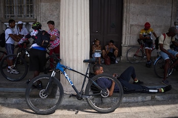 Grupo de personas con bicicletas en una calle de La Habana. Se observan hombres y mujeres de pie, sentados o recostados, con edificios y una columna detrás de ellos