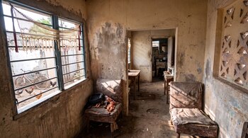 Interior de una habitación con paredes desgastadas, dos sillones viejos con muñecos de plástico, una ventana con rejas y un patio con gallinas.