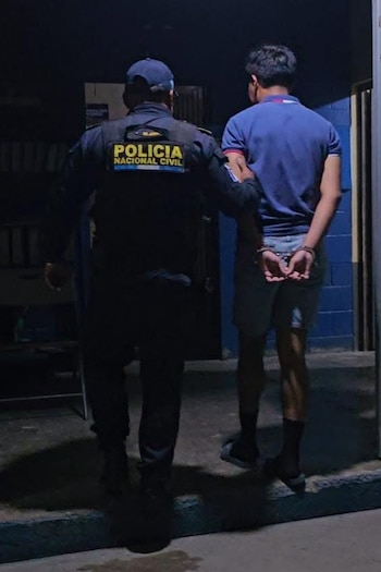 Un policía con uniforme oscuro y gorra acompaña a un hombre esposado con camisa azul y shorts, visto de espalda en un lugar con luz tenue