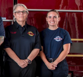 Dos mujeres uniformadas del servicio de bomberos posan de pie junto a la carrocería roja de un camión de emergencia