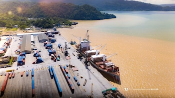 Vista aérea de un barco de carga atracado en un puerto marítimo junto a filas de contenedores y montañas al fondo