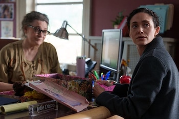 Dos mujeres en un escritorio. Una con gafas, la otra con cabello rizado sostiene papeles. Lámpara, monitor, bolígrafos, placa "Jolie Dixon Bright Fields Group Home"