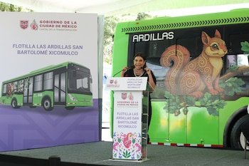 Mujer en un podio con micrófonos, un autobús verde con ilustración de ardilla y el texto ARDILLAS, y un cartel con logos y texto