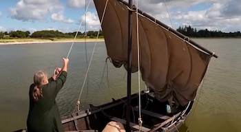 Persona de espaldas tirando de una cuerda para izar la vela marrón de un pequeño barco de madera sobre el agua
