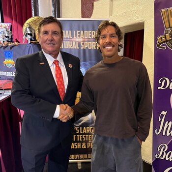 Hombre de traje y Joseph Baena, con camiseta marrón y pantalones grises, se dan la mano frente a pancartas de evento deportivo