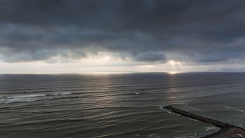Vista del mar bajo un cielo de nubes oscuras con rayos de luz solar reflejados en la superficie del agua y un espigón de piedras