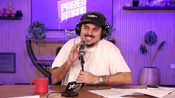 Hombre sonriente con gorra negra y auriculares, habla a un micrófono en un estudio. En el escritorio hay una taza roja y papeles; el fondo es morado con texto