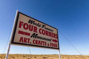 Un cartel blanco sobre postes de metal indica 'FOUR CORNERS Monument' con flechas hacia 'ART, CRAFTS & FOOD', contra un cielo azul y un paisaje desértico árido