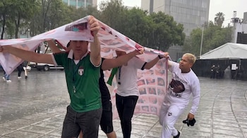 Cuatro hombres se cubren de la lluvia con una manta blanca estampada con fotografías. El suelo mojado refleja el cielo gris y edificios a lo lejos.