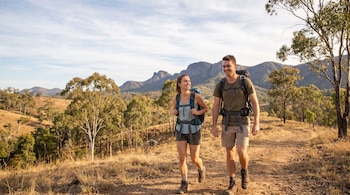 Un hombre y una mujer con mochilas caminan por un camino de tierra rodeado de vegetación y montañas al fondo.