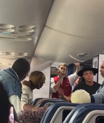 Interior de un avión de Delta con un tripulante en chaleco rojo hablando con pasajeros; una mujer y un hombre con sombrero negro son visibles en el pasillo