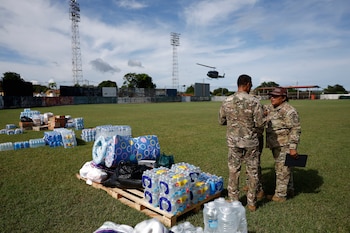 Dos militares con uniforme de camuflaje conversan en un campo junto a palés con paquetes de agua y víveres, con un helicóptero al fondo
