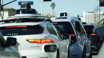 Múltiples coches Waymo blancos y azul claro con sensores en el techo circulan en fila por una calle, bajo un cielo despejado