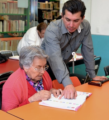 Un hombre joven con camisa a rayas apunta a un libro abierto sobre una mesa naranja, guiando a una mujer mayor con gafas y cárdigan rosa que mira el libro