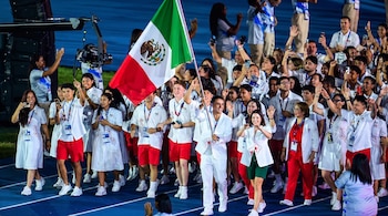 Un grupo numeroso de personas jóvenes desfila en una pista azul. Visten uniformes blancos y uno porta la bandera de México. Muchos saludan o sostienen celulares.