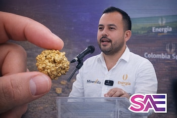 Un hombre con barba hablando ante un micrófono, un podio de acrílico transparente, una mano sosteniendo una pepita de oro, logos de Minervida, Energia y SAE.
