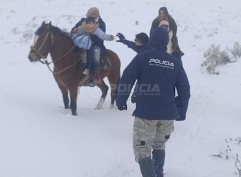 Un policía de espaldas camina por la nieve. Delante hay un caballo con un adulto y un niño. Otras dos personas están cerca del caballo en un paisaje nevado