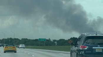 Carretera con varios vehículos, incluyendo un auto amarillo descapotable. La mediana con césped y árboles. Un letrero de salida verde. Columna de humo oscuro en el cielo