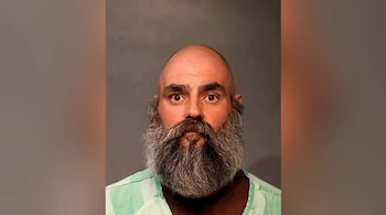 Primer plano de un hombre calvo con barba gris frondosa y camiseta verde a rayas, mirando al frente sobre un fondo gris liso