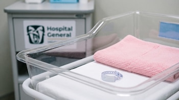 Cuna de hospital vacía con manta rosa y pulsera de identificación sobre sábana blanca. Al fondo, armario médico con el logotipo del Hospital General.