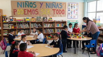 Un salón de clases de primaria con niños y una maestra, mesas redondas, sillas, estanterías con libros y juguetes, y un cartel de fomento a la igualdad.