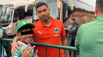 Un futbolista con camiseta naranja posa junto a un niño con gorra verde y los ojos cubiertos por una franja negra frente a un autobús.