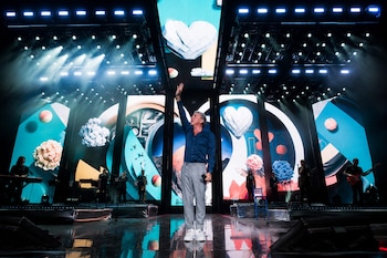 Ricardo Montaner en un escenario con luces, camisa azul y pantalón gris, saluda con el brazo levantado. Pantallas LED con formas de corazón y diseños abstractos. Músicos a los lados