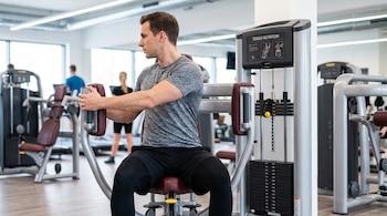 Hombre joven sentado en una máquina de rotación de torso en un gimnasio, con más personas y máquinas de ejercicio al fondo.