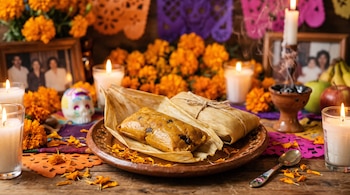 Tamales en hojas de maíz sobre un plato de barro, rodeados de velas encendidas, flores amarillas de cempasúchil, papel picado y retratos.