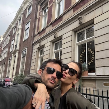 Selfie de una pareja de mediana edad, un hombre con gafas de sol abrazando a una mujer que hace un puchero, frente a un edificio de ladrillo con ventanas