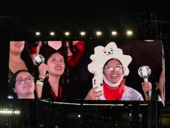 Una pantalla gigante de concierto muestra a varias personas sonrientes que sostienen objetos luminosos y llevan un gorro temático