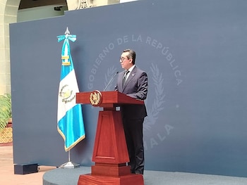 Hombre de traje y gafas en un podio con micrófono. A la izquierda, bandera de Guatemala. Fondo azul con texto 'GOBIERNO DE LA REPÚBLICA GUATEMALA'