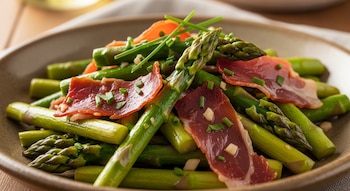Plato con espárragos verdes cocidos, lonchas de jamón serrano, trozos pequeños blancos, y cebollino picado. El plato es de color marrón oscuro.