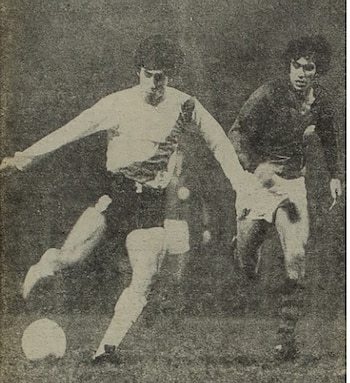 Fotografía granulada en blanco y negro de dos futbolistas en un campo de juego. Un jugador de camiseta clara patea el balón, otro de oscuro lo persigue
