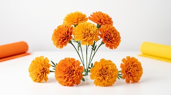 Flores de papel crepé en tonos naranja y amarillo con tallos verdes colocadas sobre una mesa blanca junto a rollos de papel.