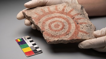 Manos con guantes sostienen un fragmento de piedra grabado con círculos rojos junto a una escala de colorimetría sobre una mesa.
