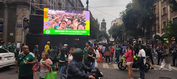 Multitud de personas en una calle con edificios, árboles, un semáforo y una pantalla gigante que proyecta una imagen de aficionados