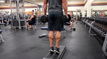 Hombre de espaldas en un gimnasio realiza ejercicio de pantorrillas sobre un step con mancuernas, sus talones están fuera del borde, otras personas y máquinas al fondo.