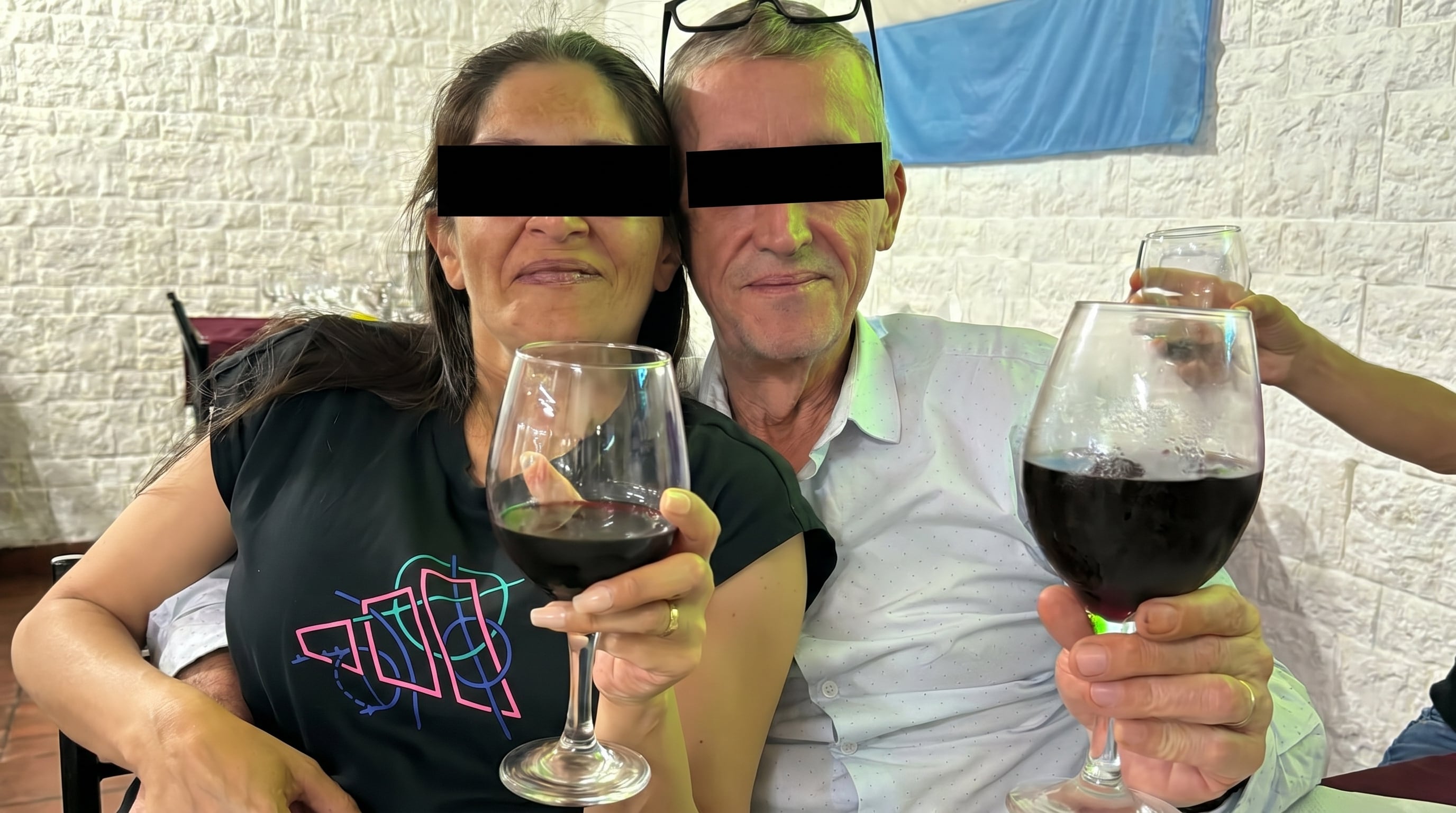 Una pareja con los ojos cubiertos con cinta negra brinda con copas de vino tinto en un ambiente festivo, con una bandera celeste y blanca de fondo. (Imagen Ilustrativa Infobae)