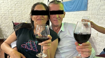 Una mujer y un hombre, con cinta negra cubriendo sus ojos, sostienen copas de vino tinto en primer plano, con una bandera celeste y blanca de fondo.