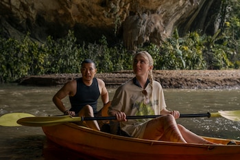 Dos personas en un kayak amarillo, con remo, en un río. La mujer tiene marcas rojas en la pierna. Al fondo hay una cueva rocosa con vegetación