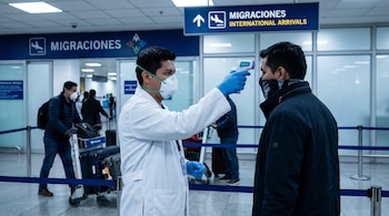 Un trabajador sanitario con bata blanca, mascarilla y guantes azules, mide la temperatura con un termómetro digital a un viajero en la zona de llegadas de un aeropuerto.