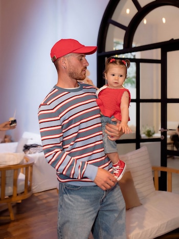Un hombre con gorra roja y camiseta a rayas sostiene en brazos a una niña pequeña vestida de rojo