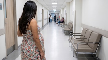 Vista trasera de una mujer latina embarazada con vestido floral en un pasillo de hospital, con sillas de espera beige y otras personas al fondo.