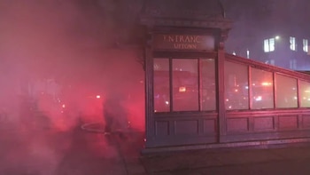 Entrada de un edificio con paneles de cristal y el letrero 'Entrance Uptown', parcialmente cubierta por humo rojo y reflejos de luces en la noche