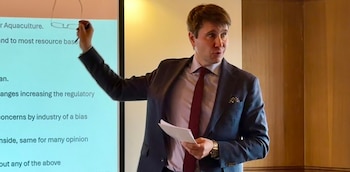 Un hombre caucásico en traje y corbata roja señala una pantalla de proyección con texto, mientras sujeta gafas y documentos en la otra mano