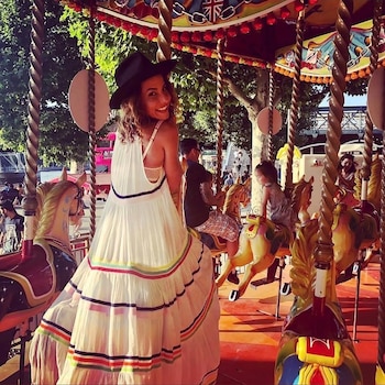 Una mujer sonriente con sombrero oscuro y vestido blanco con rayas de colores posa de espaldas en un carrusel, con otros jinetes y árboles de fondo
